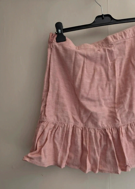 A light pink linen-textured skirt with a ruffled hem, hanging from a black plastic hanger against a plain beige wall.