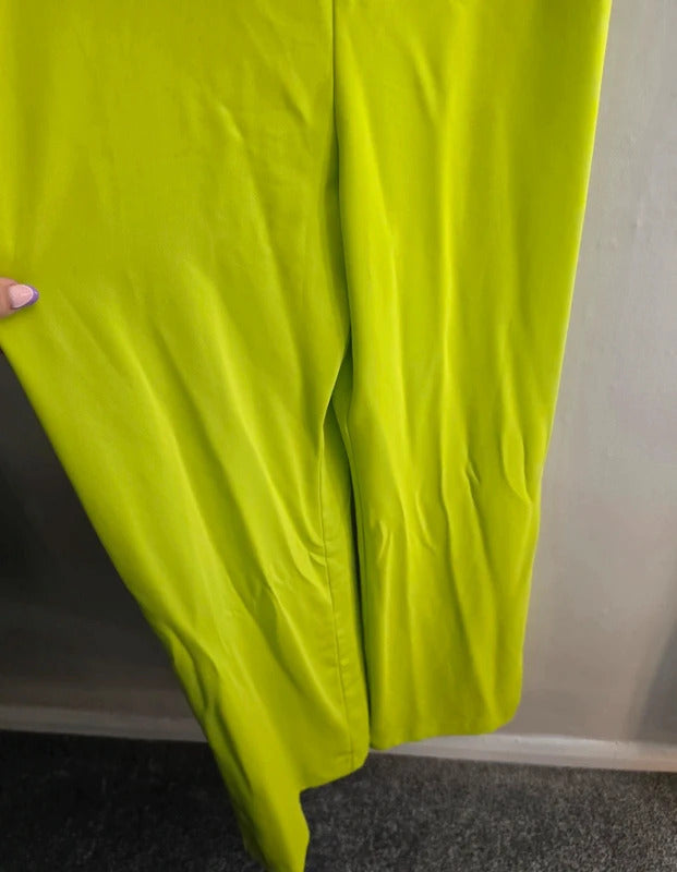 A close-up of bright lime green pants being held up by a hand, showing the fabric texture and folds against a neutral indoor background.