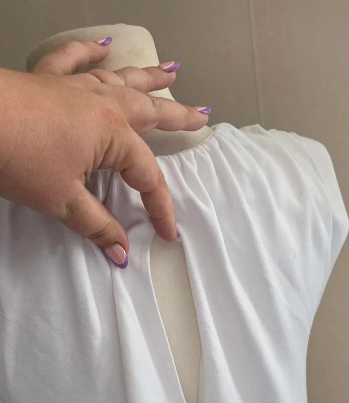 A close-up of a white sleeveless dress displayed on a mannequin torso, with a hand pointing to the gathered neckline and front opening. The mannequin is indoors against a plain beige wall.