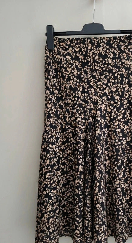 A black midi skirt with a dense beige and pink floral print, hanging on a black hanger against a plain off-white wall.