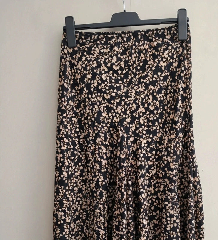 A black midi skirt with a dense beige floral print, hanging on a black hanger against a plain off-white wall.