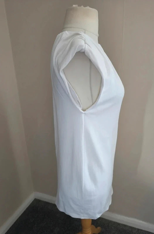 A white sleeveless dresd with a large draped shoulder cutout, displayed on a dress form mannequin in an indoor corner setting.